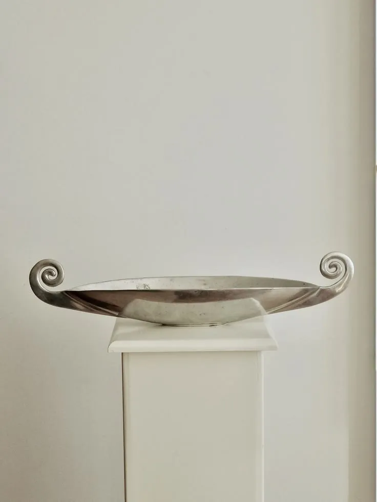 Shallow silver dish with spiral scroll handles on a white plinth